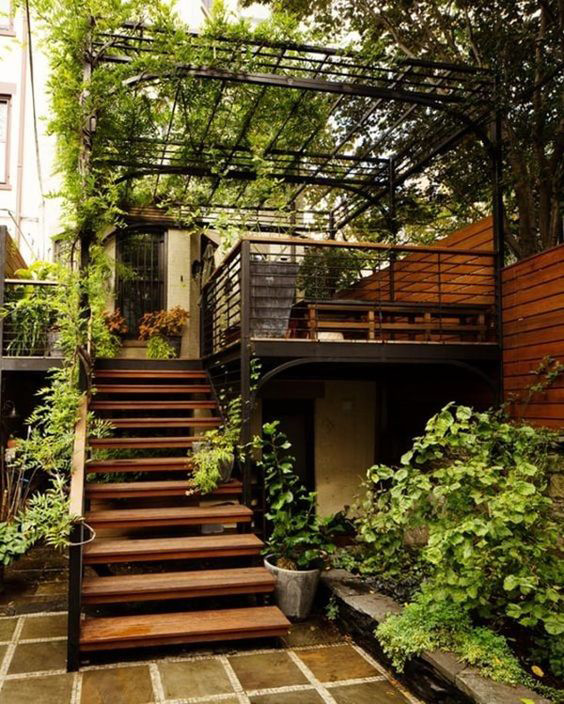 Escalera exterior de madera con pérgola metálica cubierta de plantas en un patio ajardinado.