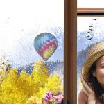 Publicidad de la campaña 'Asómate a una ventana infinita' de Saint-Gobain y ASOMA, mostrando una ventana de madera con vidrio Climalit Oraé, y en el reflejo, una madre y su hija sonriendo con sombreros frente a un paisaje con montañas, árboles amarillos, un globo aerostático y pájaros volando.